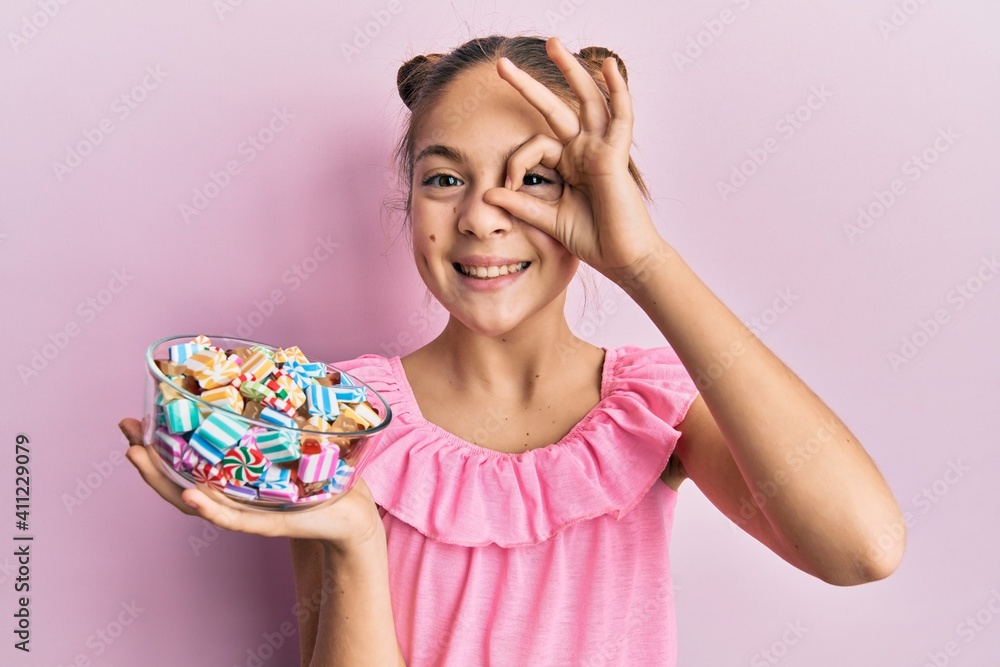 Beautiful brunette little girl holding jar full of sugar candies smiling happy doing ok sign with hand on eye looking through fingers Beautiful brunette little girl holding jar full of sugar candies smiling happy doing ok sign with hand on eye looking through fingers