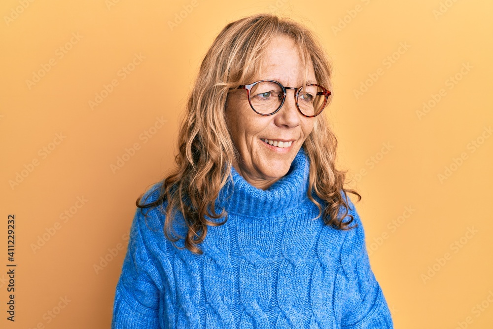 Middle age blonde woman wearing glasses and casual winter sweater looking away to side with smile on face, natural expression. laughing confident. Middle age blonde woman wearing glasses and casual winter sweater looking away to side with smile on face, natural expression. laughing confident.
