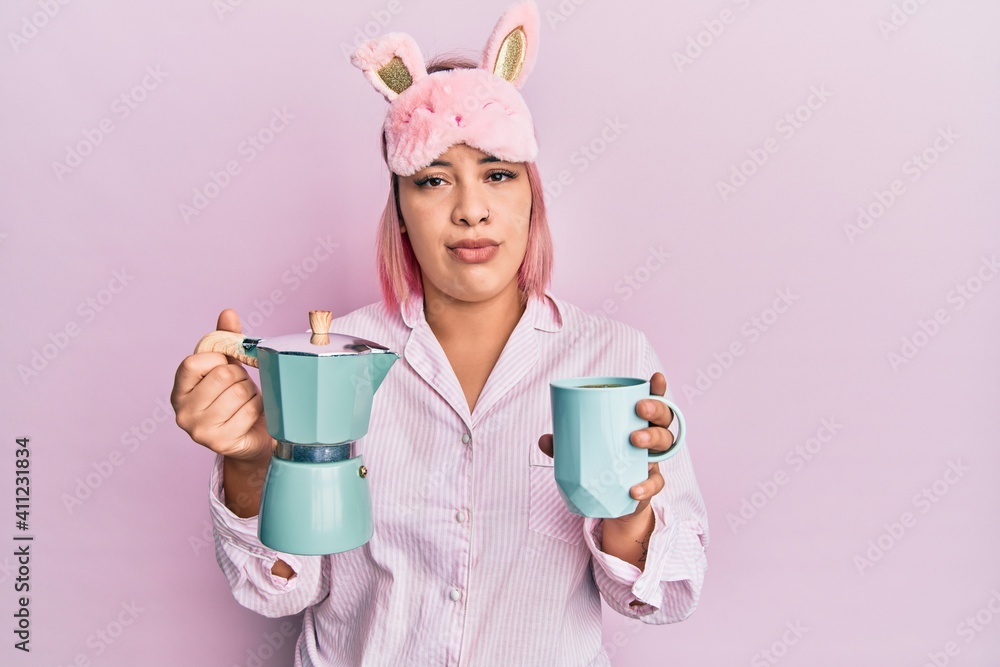 Hispanic woman with pink hair wearing sleep mask and pajama drinking a coffee cup relaxed with serious expression on face. simple and natural looking at the camera. Hispanic woman with pink hair wearing sleep mask and pajama drinking a coffee cup relaxed with serious expression on face. simple and natural looking at the camera.
