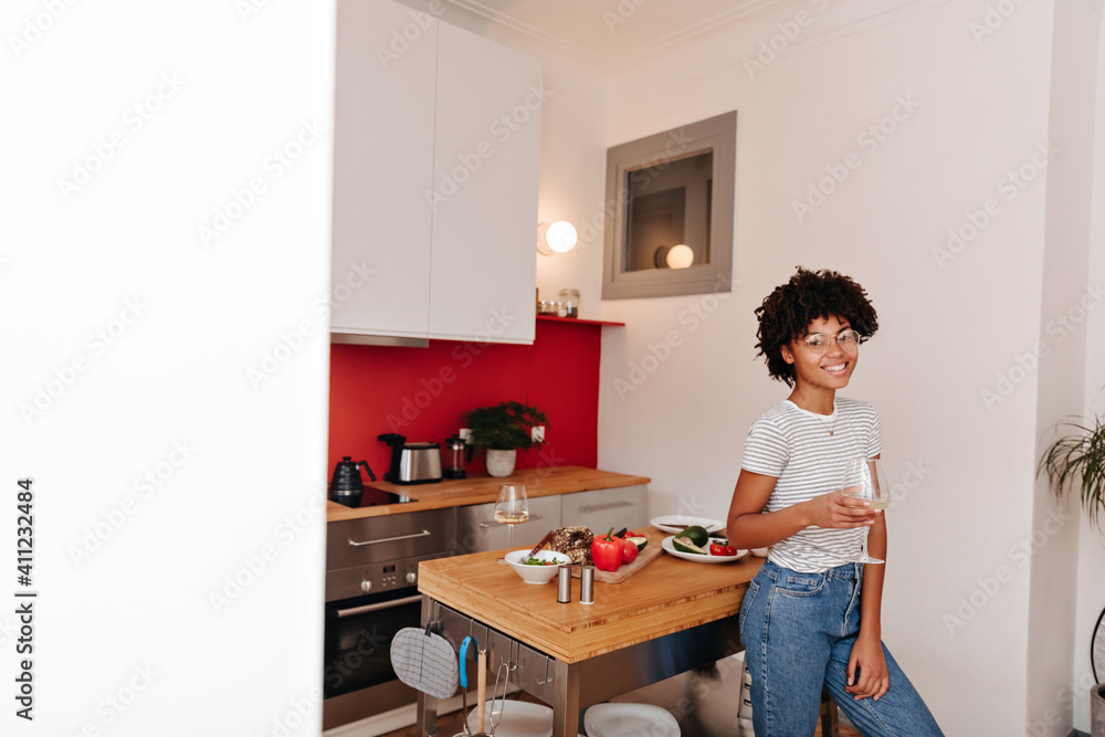 Beautiful curly girl in jeans leans on kitchen table with vegetables and holds glass of wine Beautiful curly girl in jeans leans on kitchen table with vegetables and holds glass of wine