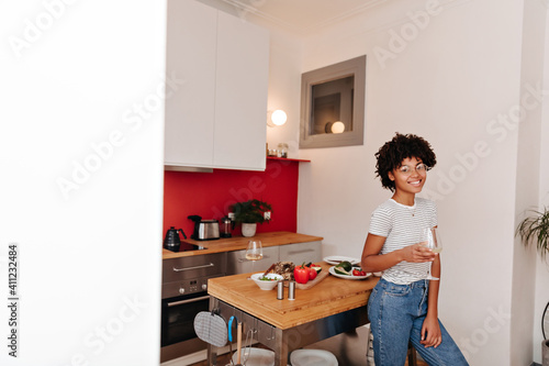 Beautiful curly girl in jeans leans on kitchen table with vegetables and holds glass of wine