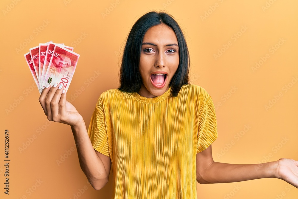 Young brunette woman holding 20 israel shekels banknotes celebrating achievement with happy smile and winner expression with raised hand Young brunette woman holding 20 israel shekels banknotes celebrating achievement with happy smile and winner expression with raised hand