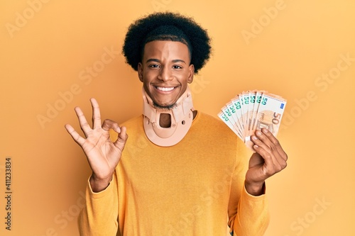 African american man with afro hair wearing cervical neck collar and holding money from insurance doing ok sign with fingers, smiling friendly gesturing excellent symbol