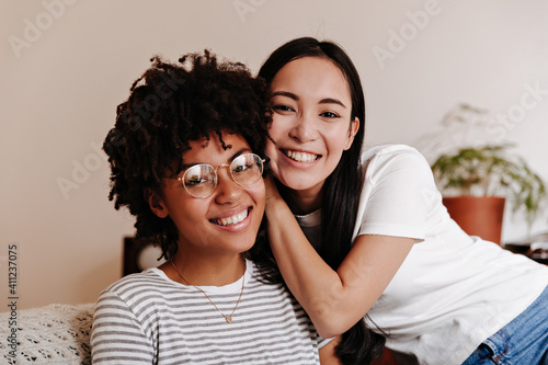 Portrait of dark-skinned curly woman in glasses and her Asian long-haired girlfriend in white T-shirt