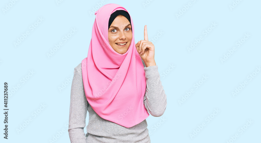 Young caucasian woman wearing traditional islamic hijab scarf pointing finger up with successful idea. exited and happy. number one.