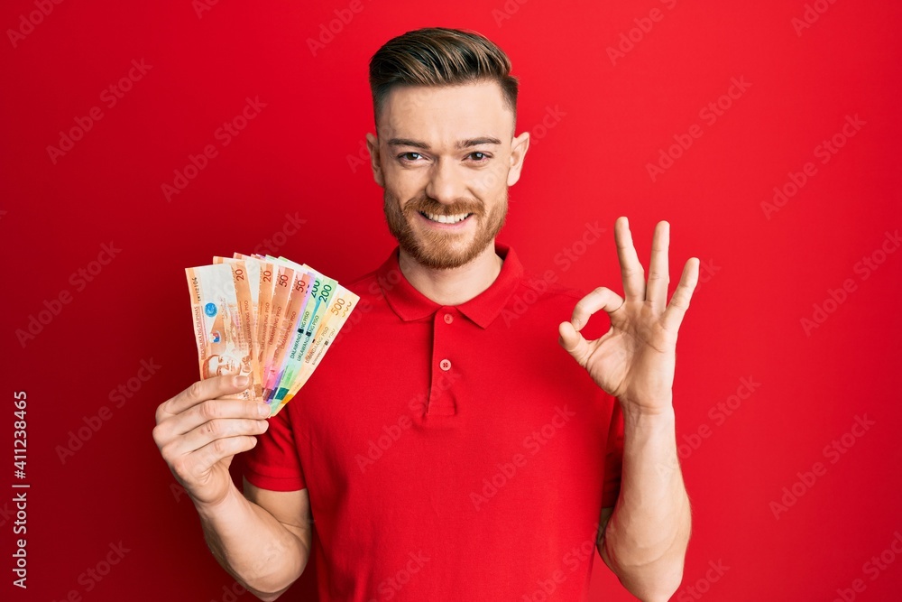Young redhead man holding philippine peso banknotes doing ok sign with fingers, smiling friendly gesturing excellent symbol Young redhead man holding philippine peso banknotes doing ok sign with fingers, smiling friendly gesturing excellent symbol