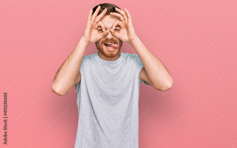 Young redhead man wearing casual grey t shirt doing ok gesture like binoculars sticking tongue out, eyes looking through fingers. crazy expression. Young redhead man wearing casual grey t shirt doing ok gesture like binoculars sticking tongue out, eyes looking through fingers. crazy expression.