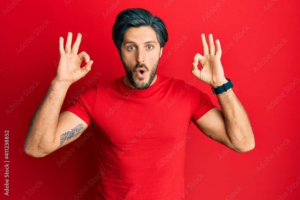 Young hispanic man wearing casual red t shirt looking surprised and shocked doing ok approval symbol with fingers. crazy expression Young hispanic man wearing casual red t shirt looking surprised and shocked doing ok approval symbol with fingers. crazy expression