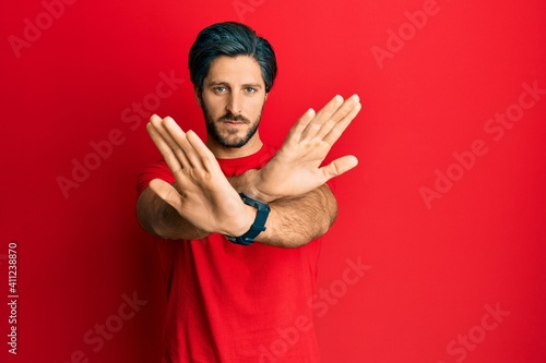 Young hispanic man wearing casual red t shirt rejection expression crossing arms and palms doing negative sign, angry face