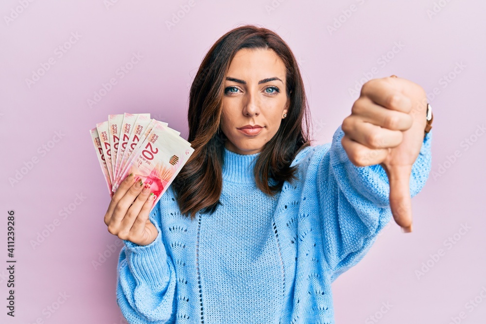 Young brunette woman holding 20 israel shekels banknotes looking unhappy and angry showing rejection and negative with thumbs down gesture. bad expression. Young brunette woman holding 20 israel shekels banknotes looking unhappy and angry showing rejection and negative with thumbs down gesture. bad expression.
