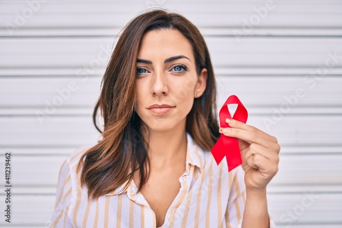 Картината върху платно Young hispanic woman with serious expression holding red hiv ribbon standing at the city