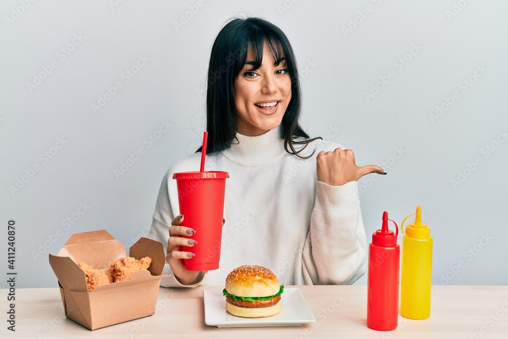 Young brunette woman with bangs eating a tasty classic burger with ketchup and mustard pointing thumb up to the side smiling happy with open mouth Young brunette woman with bangs eating a tasty classic burger with ketchup and mustard pointing thumb up to the side smiling happy with open mouth