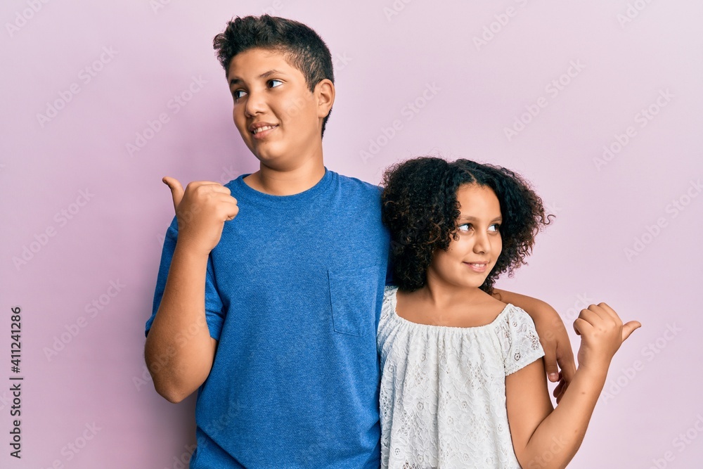 Young hispanic family of brother and sister wearing casual clothes together smiling with happy face looking and pointing to the side with thumb up. Young hispanic family of brother and sister wearing casual clothes together smiling with happy face looking and pointing to the side with thumb up.