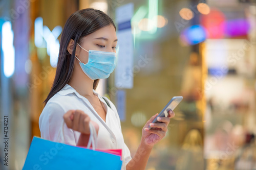 The life during outbreak of Covid-19 concept: Young Asian female with face mask use smart phone in shopping mall.