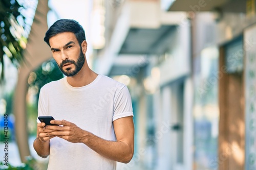 Young hispanic man with serious expression using smartphone at the city.