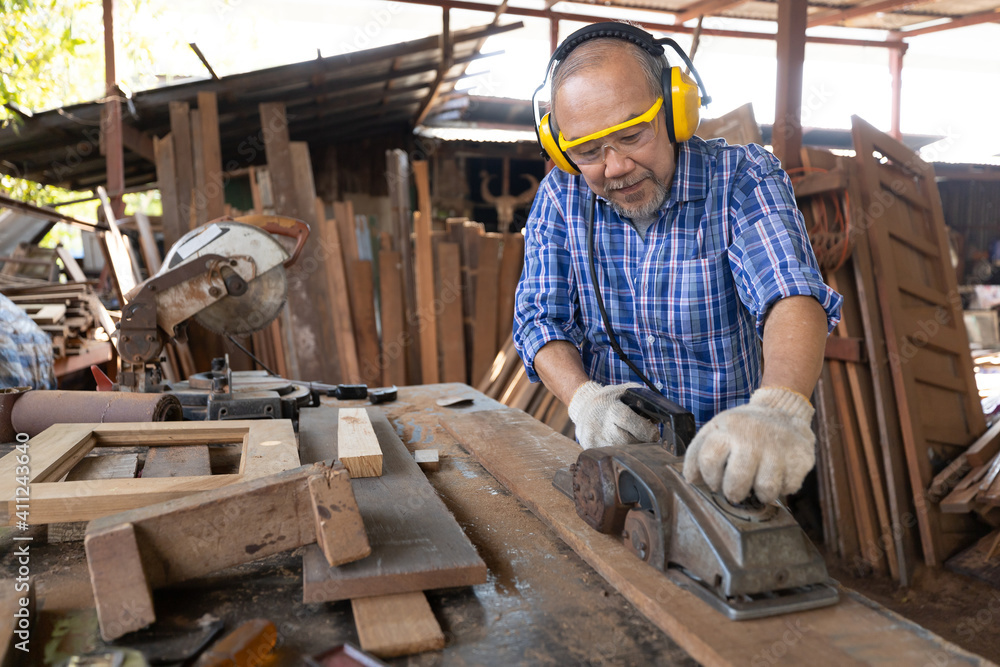 senior asian man carpenter wearing glasses and headphone, using electric wood planers on a piece of wood in workshop - obrazy, fototapety, plakaty 