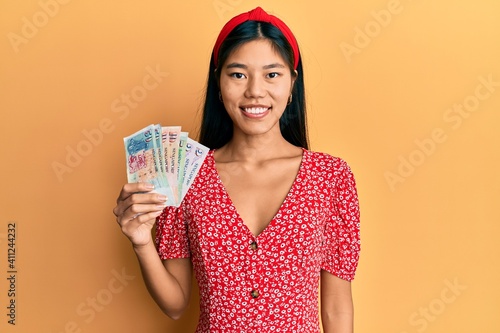 Photography Young chinese woman holding singapore dollars banknotes looking positive and hap