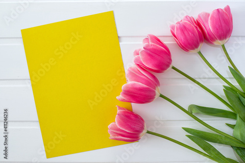Purple-pink tulips and a yellow sheet of paper on a white background, spring and Easter card, top view, space for text