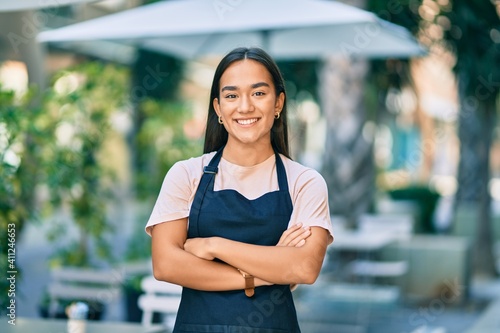 Young latin barista girl with arms crossed smiling happy at the coffee shop