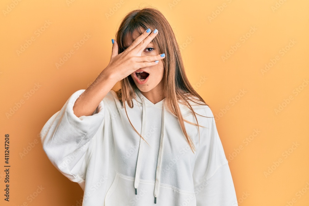 Teenager caucasian girl wearing casual sweatshirt peeking in shock covering face and eyes with hand, looking through fingers afraid Teenager caucasian girl wearing casual sweatshirt peeking in shock covering face and eyes with hand, looking through fingers afraid