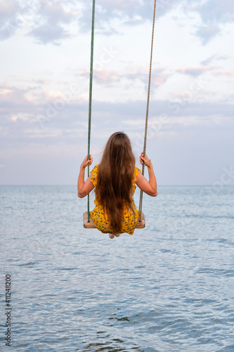 Girl with beautiful long hair sits on swing above sea. Back view. Vertical frame.