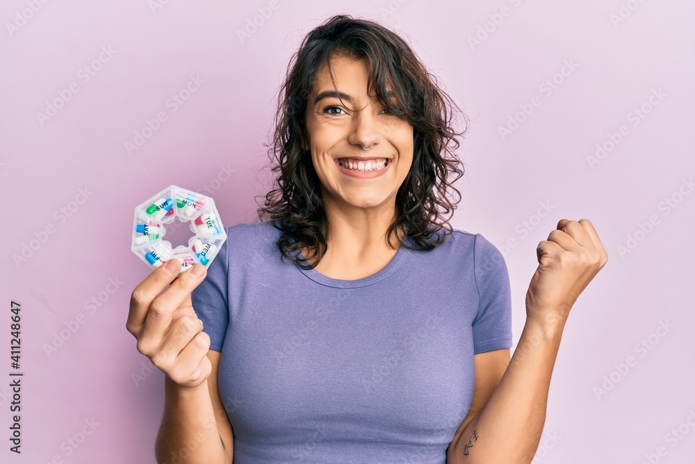 Young hispanic woman holding pill organizer screaming proud, celebrating victory and success very excited with raised arm Young hispanic woman holding pill organizer screaming proud, celebrating victory and success very excited with raised arm