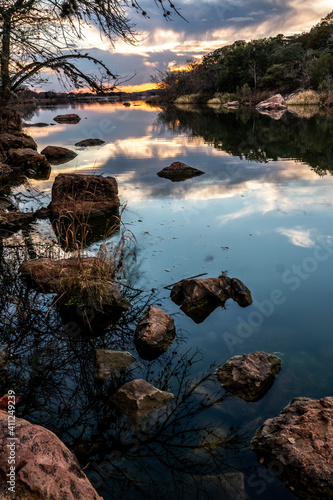 Rocks protrude through the perfectly calm surface of a lake with reflection of the sun and clouds at sunset, Inks Lake, Texas
