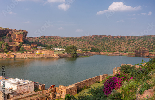 Badami, Karnataka, India - November 7, 2013: Cave temples above Agasthya Lake in NE-view, surrounded by hills, rocks, and cliffs under blue cloudscape. Flowers add color, Green foliage.