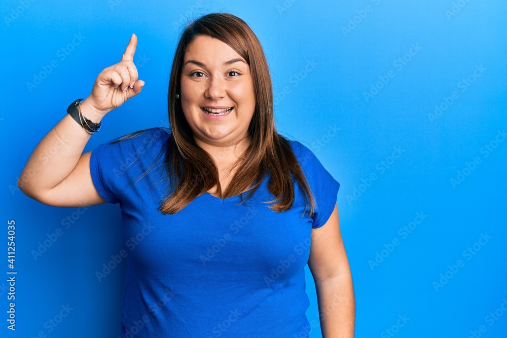 Beautiful brunette plus size woman wearing casual blue t shirt pointing finger up with successful idea. exited and happy. number one. Beautiful brunette plus size woman wearing casual blue t shirt pointing finger up with successful idea. exited and happy. number one.