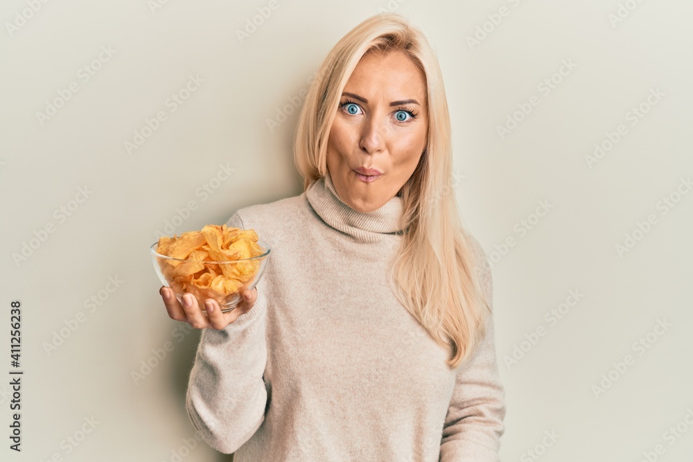 Young blonde woman holding bowl with potato chip scared and amazed with open mouth for surprise, disbelief face