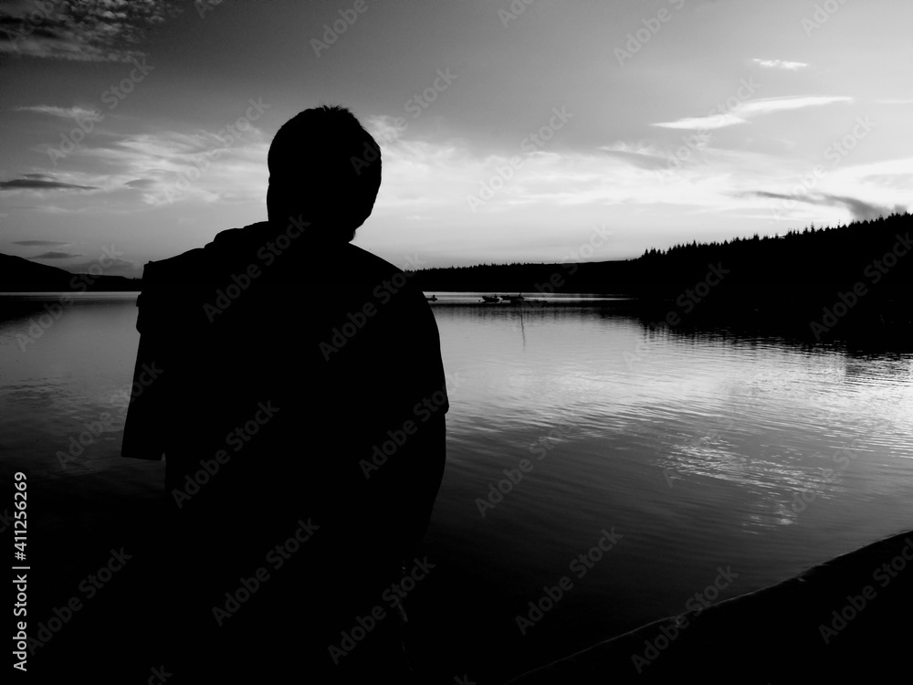 Fototapeta premium Black and white silhouette of a person on lake Loch Lomond during sunset with some clouds and a background forest
