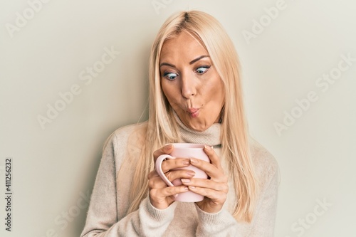 Young blonde woman drinking a cup of coffee making fish face with mouth and squinting eyes  crazy and comical.