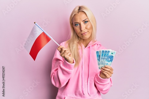 Obraz na plátně Young blonde woman holding poland flag and zloty banknotes relaxed with serious expression on face