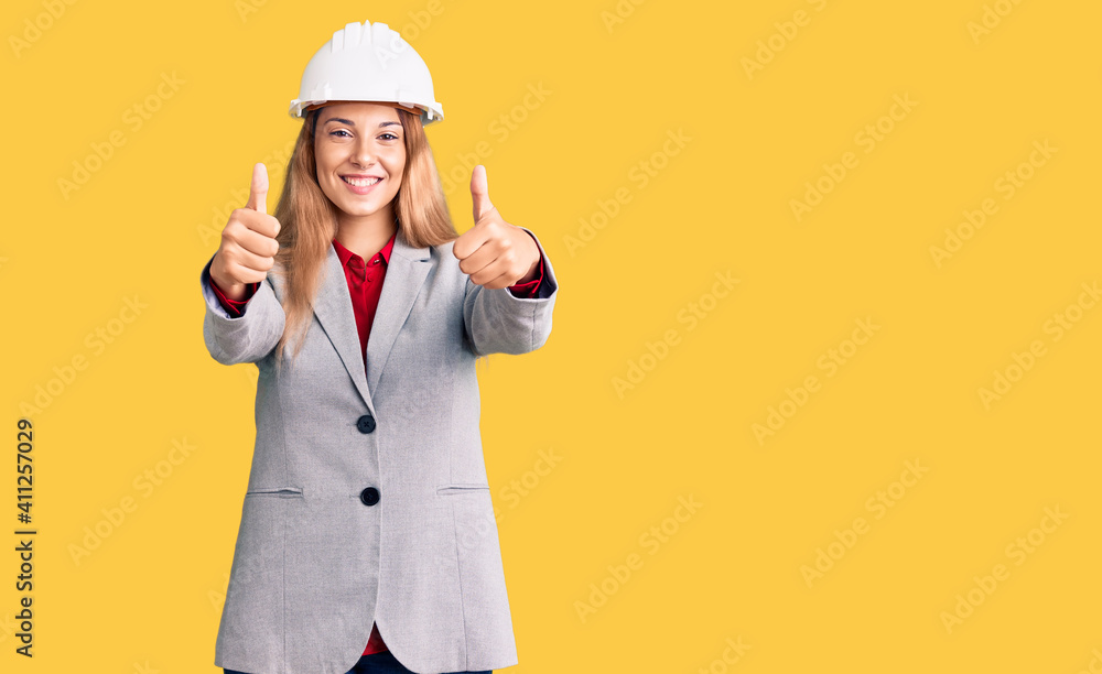 Beautiful young woman wearing architect hardhat approving doing positive gesture with hand, thumbs up smiling and happy for success. winner gesture.
