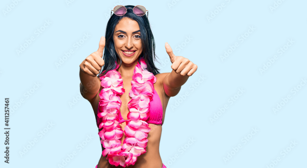 Beautiful hispanic woman wearing bikini and hawaiian lei approving doing positive gesture with hand, thumbs up smiling and happy for success. winner gesture. Beautiful hispanic woman wearing bikini and hawaiian lei approving doing positive gesture with hand, thumbs up smiling and happy for success. winner gesture.