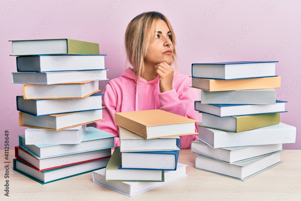 Young caucasian woman sitting on the table with books with hand on chin thinking about question, pensive expression. smiling with thoughtful face. doubt concept. Young caucasian woman sitting on the table with books with hand on chin thinking about question, pensive expression. smiling with thoughtful face. doubt concept.