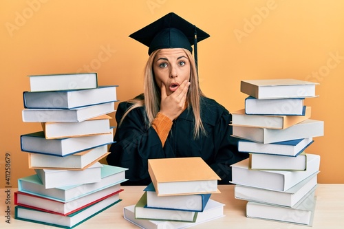 Young caucasian woman wearing graduation ceremony robe sitting on the table looking fascinated with disbelief, surprise and amazed expression with hands on chin