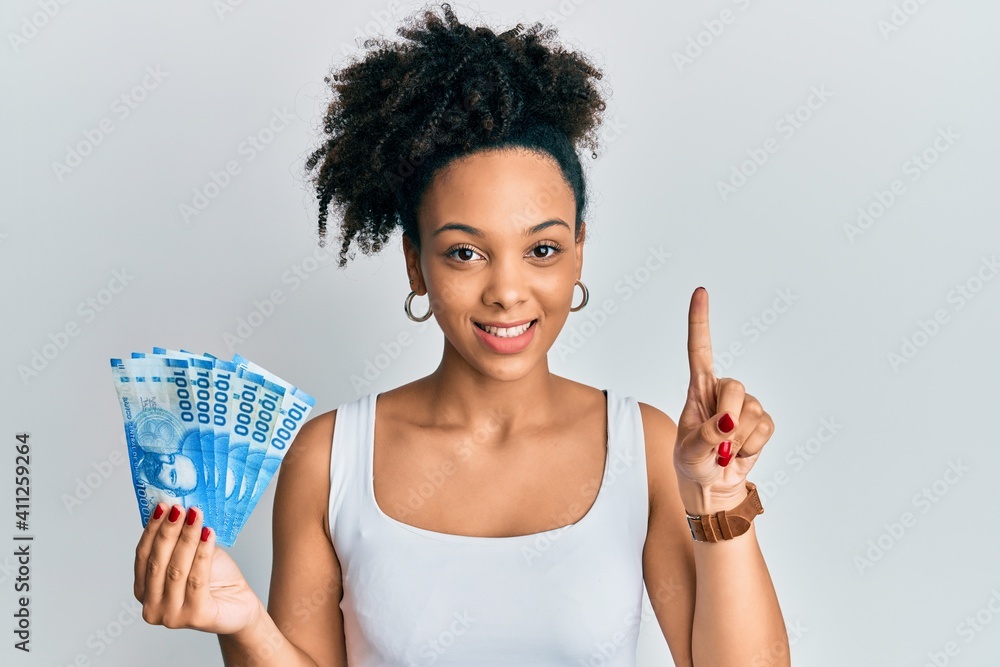 Young african american girl holding 10000 chilean pesos smiling with an idea or question pointing finger with happy face, number one Young african american girl holding 10000 chilean pesos smiling with an idea or question pointing finger with happy face, number one