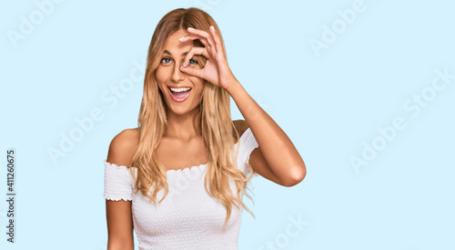Beautiful blonde young woman wearing casual white tshirt doing ok gesture with hand smiling, eye looking through fingers with happy face.