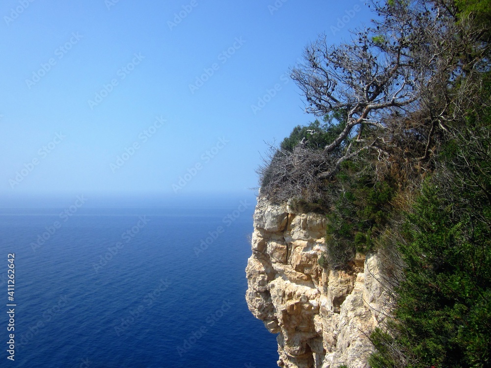 Hanging cliff rock with a pine tree against a blue sea water and blue sky. Beautiful background, marine and holiday concept, perfect  for motivation quote. Zakynthos. Greece. Free space for text