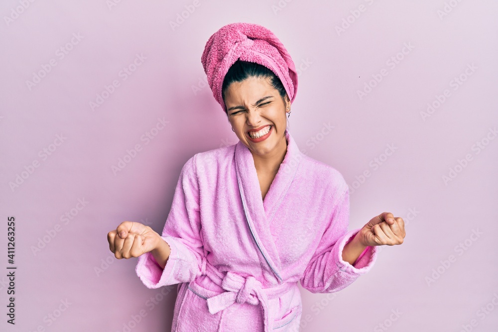 Young hispanic woman wearing shower towel cap and bathrobe very happy and excited doing winner gesture with arms raised, smiling and screaming for success. celebration concept. Young hispanic woman wearing shower towel cap and bathrobe very happy and excited doing winner gesture with arms raised, smiling and screaming for success. celebration concept.