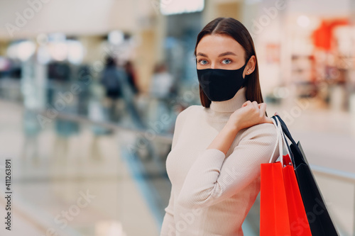 Young adult woman wearing black protective medical face mask carrying paper shopping bags in hands at mall. Black friday and covid-19 new normal.