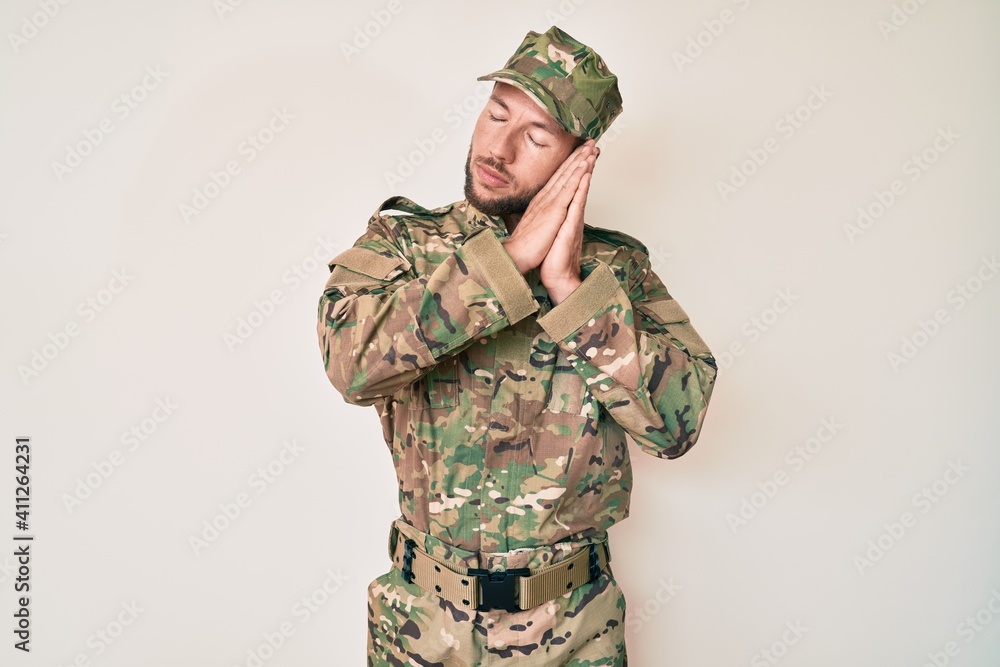 Young caucasian man wearing camouflage army uniform sleeping tired dreaming and posing with hands together while smiling with closed eyes. Young caucasian man wearing camouflage army uniform sleeping tired dreaming and posing with hands together while smiling with closed eyes.