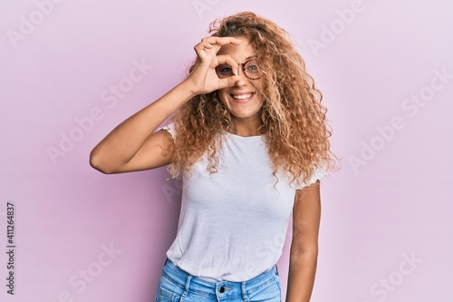Beautiful caucasian teenager girl wearing white t-shirt over pink background doing ok gesture with hand smiling, eye looking through fingers with happy face.