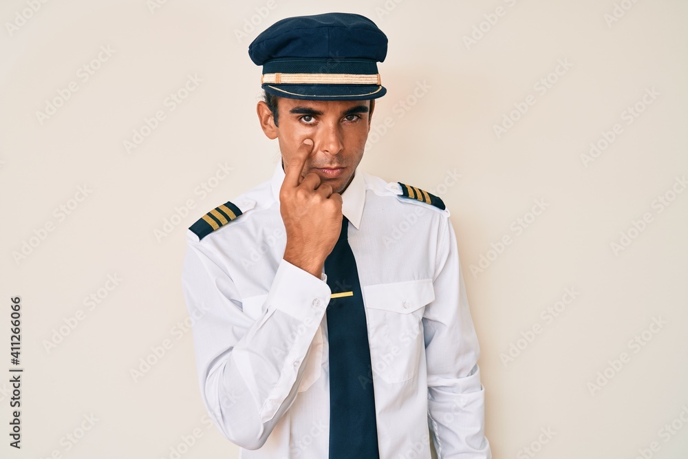 Young hispanic man wearing airplane pilot uniform pointing to the eye watching you gesture, suspicious expression Young hispanic man wearing airplane pilot uniform pointing to the eye watching you gesture, suspicious expression