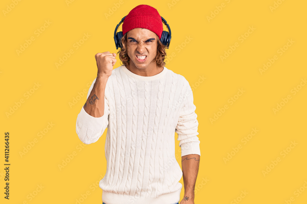 Fototapeta premium Young hispanic man listening to music using headphones angry and mad raising fist frustrated and furious while shouting with anger. rage and aggressive concept.