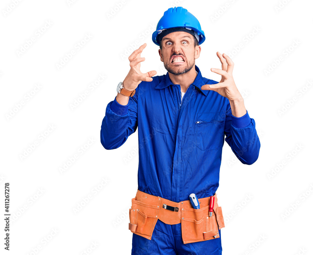Young handsome man wearing worker uniform and hardhat angry and mad raising fist frustrated and furious while shouting with anger. rage and aggressive concept.