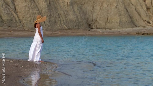 Woman alone in a white dress and hat standing in front of the sea on a deserted beach thinking, nostalgia, emotions, memories 