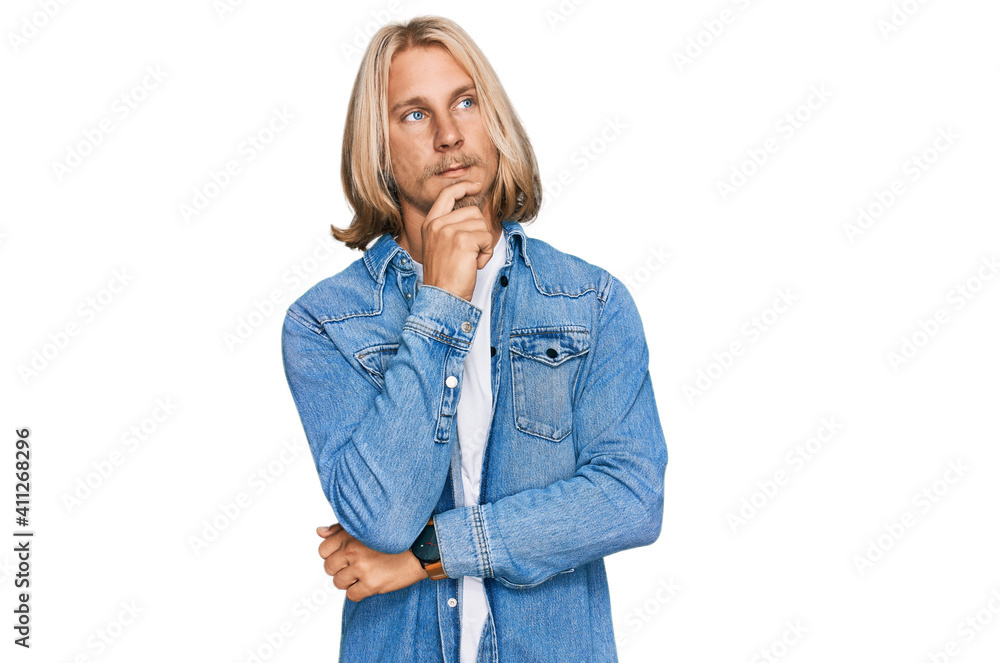 Caucasian man with blond long hair wearing casual denim jacket with hand on chin thinking about question, pensive expression. smiling with thoughtful face. doubt concept. Caucasian man with blond long hair wearing casual denim jacket with hand on chin thinking about question, pensive expression. smiling with thoughtful face. doubt concept.