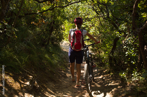 Tired woman with backpack and helmet walking next to her bicycle in the forest path of Strunjan